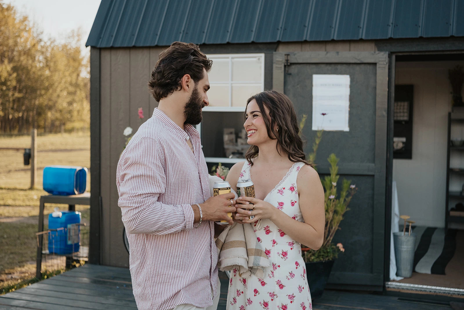 Man and woman on a date night standing in front of a small building at Blooms on 7 flower farm, holding a local Alberta craft beer and smiling.
