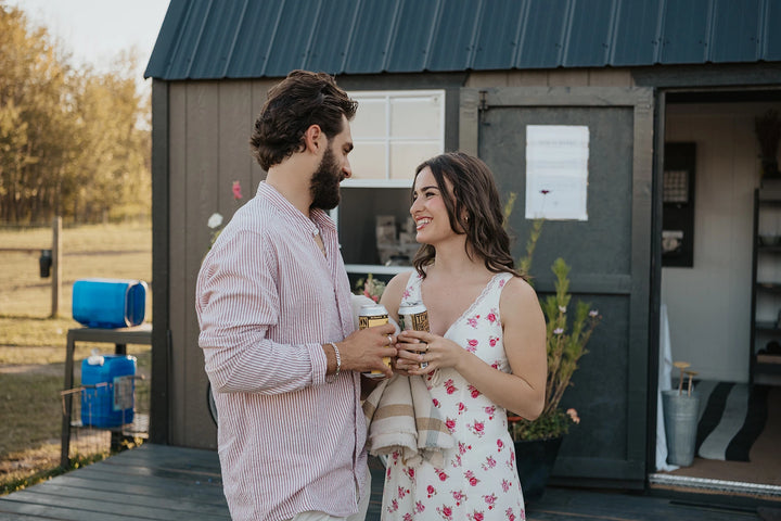 Man and woman on a date night standing in front of a small building at Blooms on 7 flower farm, holding a local Alberta craft beer and smiling.
